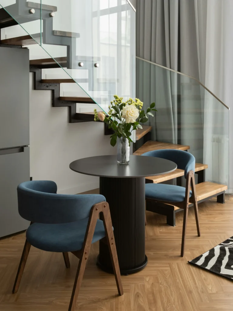 Rincón moderno con mesa redonda, sillas tapizadas y escalera de madera en interior contemporáneo.
